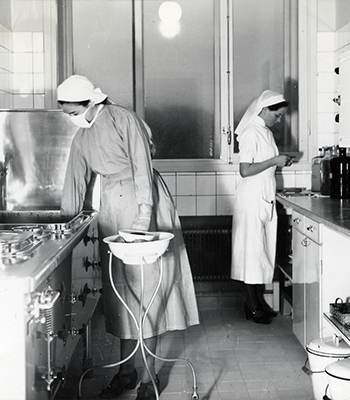 Historiskt svartvitt foto av två vårdklädda kvinnor i sjukhusmiljö.
