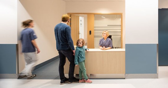 Pappa med hans barn står framför en receptionsdisk på ett sjukhus. Bakom sitter en vårdpersonal och tar emot dem.