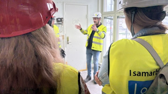 En från Byggdialog har rundvisning av IVA/MIVA projektet. En man i gul varsel väst står och berättar för en publik med gula västar.