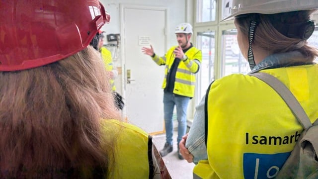 En från Byggdialog har rundvisning av IVA/MIVA projektet. En man i gul varsel väst står och berättar för en publik med gula västar.