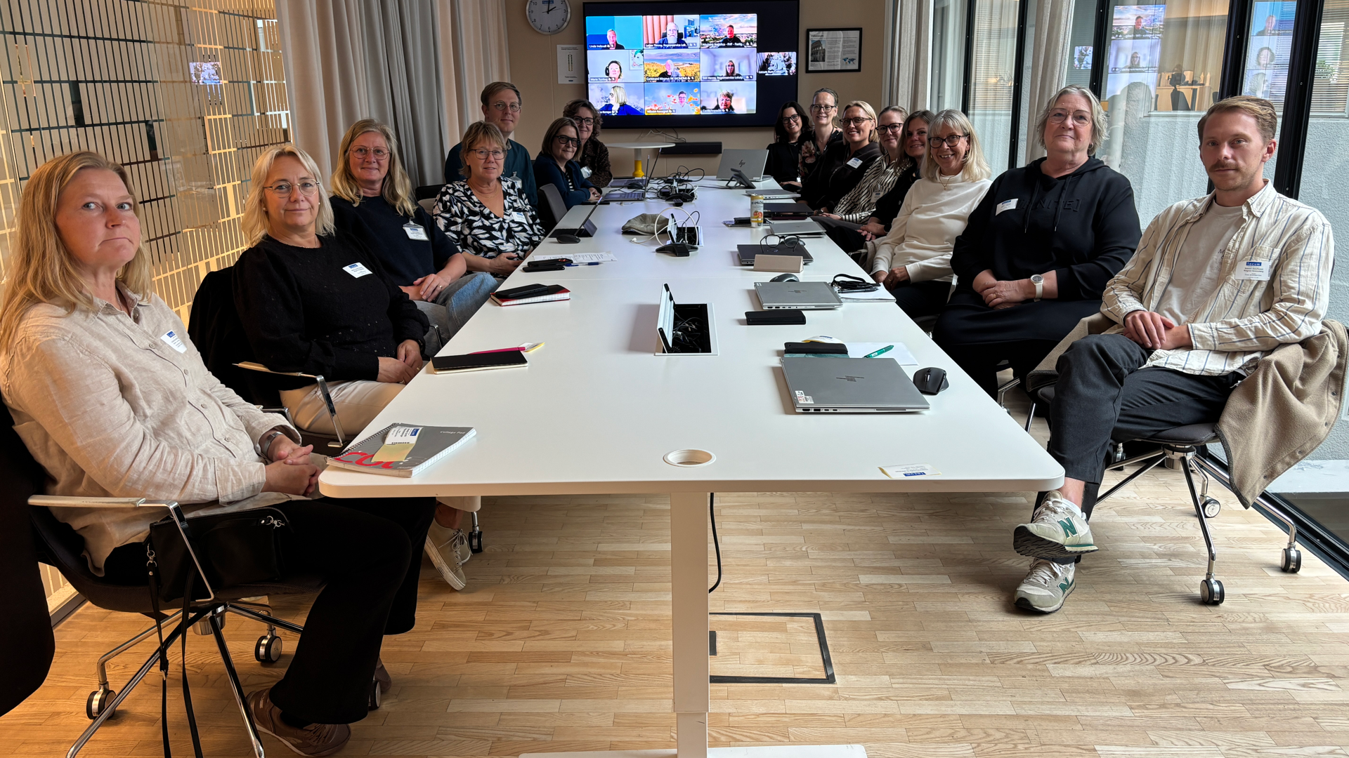 En grupp personer sitter runt ett avl&aring;ngt, vitt konferensbord med n&aring;gra personer p&aring; bild l&auml;ngst fram p&aring; en sk&auml;rm.