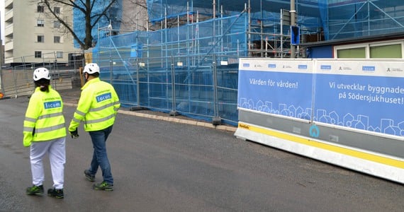 En man och en kvinna i gula varseljackor och vit bygghjälm går förbi utanför byggområdet.