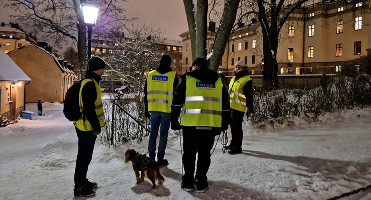 En grupp män som står bredvid en hund på en snötäckt gata med gula reflexväster. De går en trygghetsrondering på Landstingstomten.