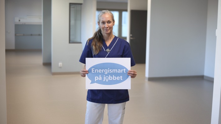 En vårdpersonal håller en skylt i ett tomt rum. Texten på skylten är