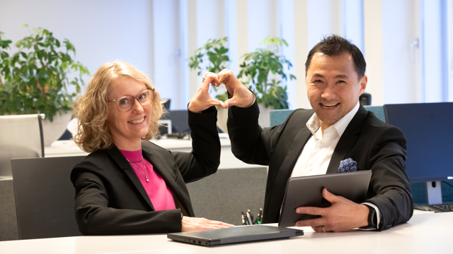 Två av Locums chefer står vid ett bord med en bärbar dator och gör hjärttecken med händer.