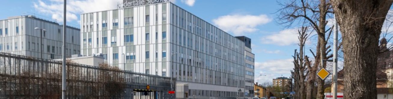 En bil som kör längs en gata bredvid en hög byggnad. På taket sitter en skylt där det står Södertälje sjukhus