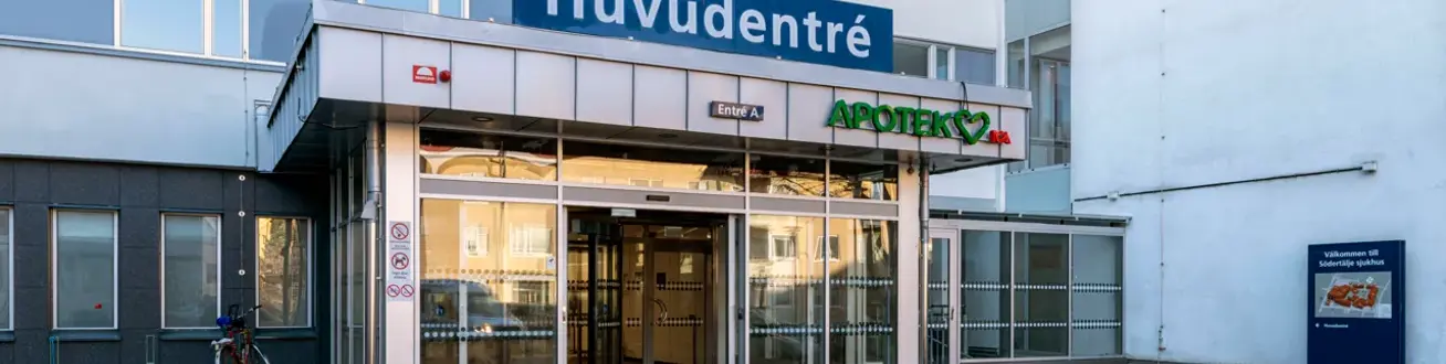 Fasaden på hus 01 med huvudentrén. Skjutdörrarna står öppna och solen skiner på glaspartierna.
