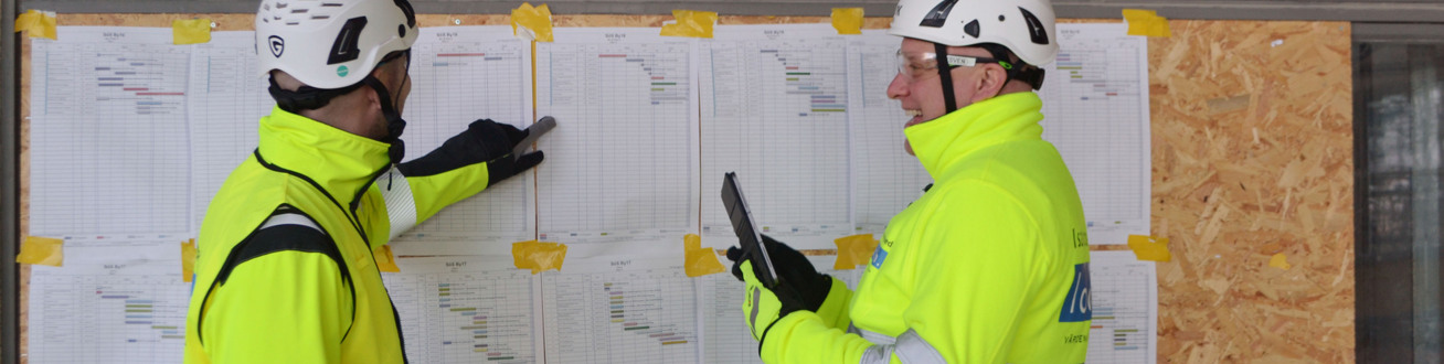 Två personer med hjälm och varseljacka pratar och tittar på listor.