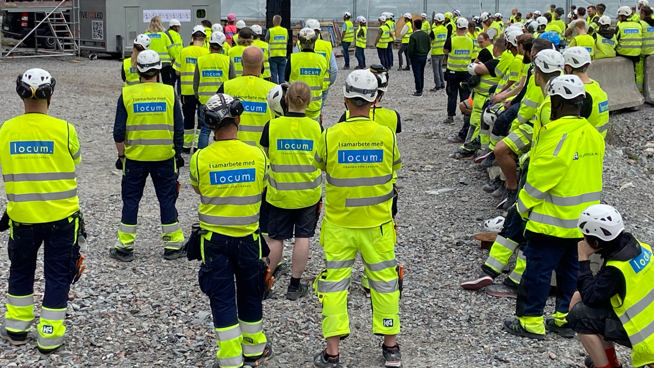 Stor grupp byggarbetare med gula varselvästar och hjälmar på grusplan, fotade bakifrån.