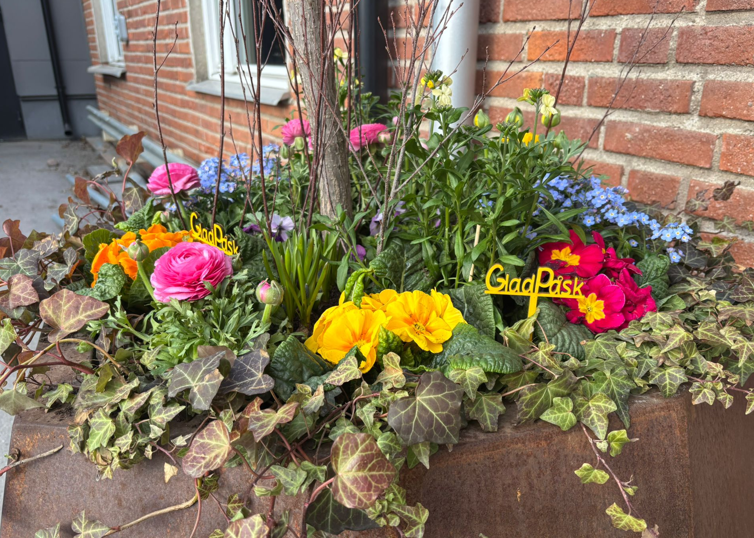 En kruka fylld med färgglada blommor bredvid en tegelbyggnad. Skylt med glad Påsk