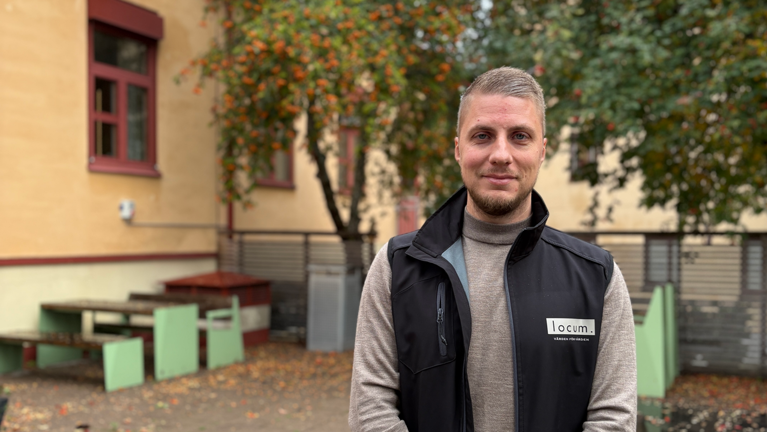 Man i svart väst står framför en byggnad och ler.