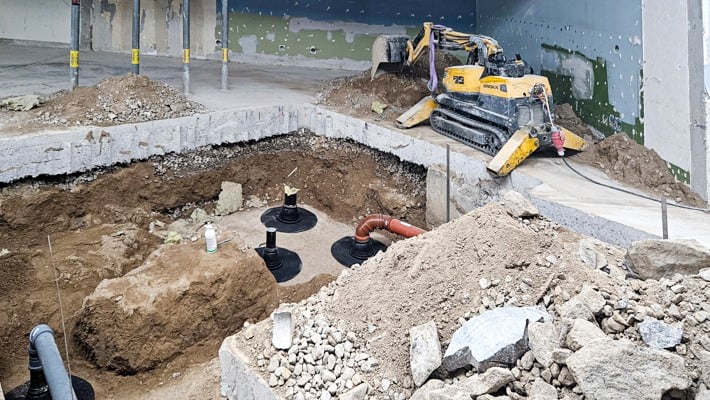 Ett stort hål i golvet där avloppsstammarna syns. I bakgrunden en liten bulldozer.