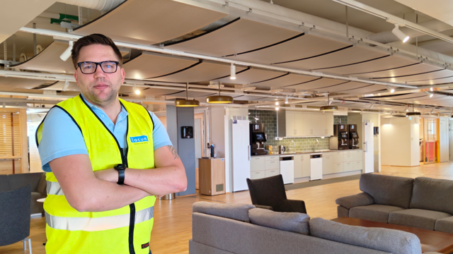 En man i gul väst står i ett stort rum med kök och soffgrupper.