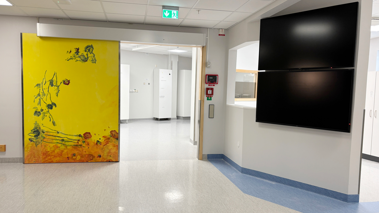 Skjutd&ouml;rr med f&auml;rgglad, gul konst i ljus v&aring;rdlokal med stor TV-sk&auml;rm p&aring; v&auml;ggen.