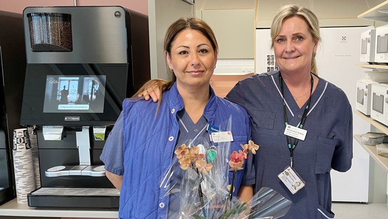 Två kvinnor i vårdkläder framför kaffeautmat med blomma i handen.