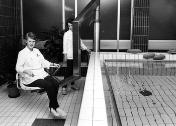 Personal sitter i stolshiss vid en rehabiliteringsbassäng. En annan kvinna styr stolshissen.