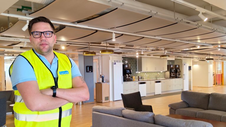 En man i gul väst står i ett stort rum med kök och soffgrupper.