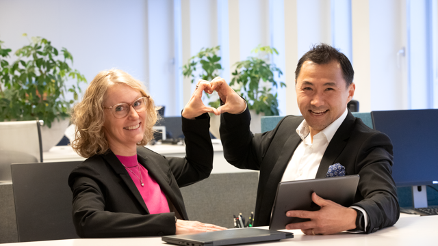 Två av Locums chefer står vid ett bord med en bärbar dator och gör hjärttecken med händer.