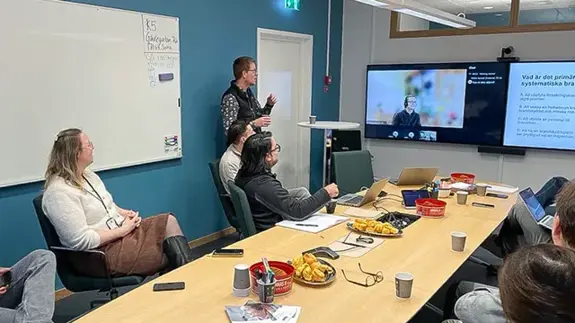 Flera personer sitter runt ett bord och tittar på en skärm. 