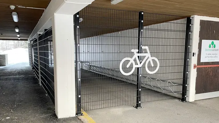 Inhägnad cykelparkering med vit cykelsymbol på väggens galler.