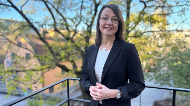 Kvinna i svart kavaj och glasögon framför svart järnstaket med grönska bakom.