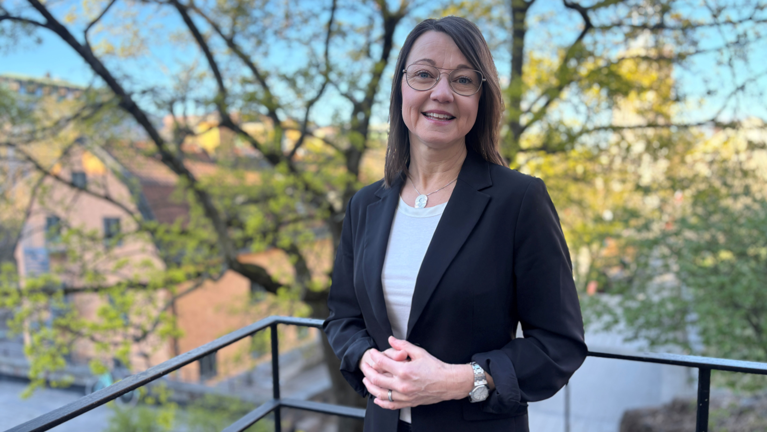 Kvinna i svart kavaj och glasögon framför svart järnstaket med grönska bakom.