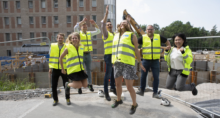 Delar av projektledningen från projekt ny vårdbyggnad hoppar i gula västar vid byggarbetsplats.