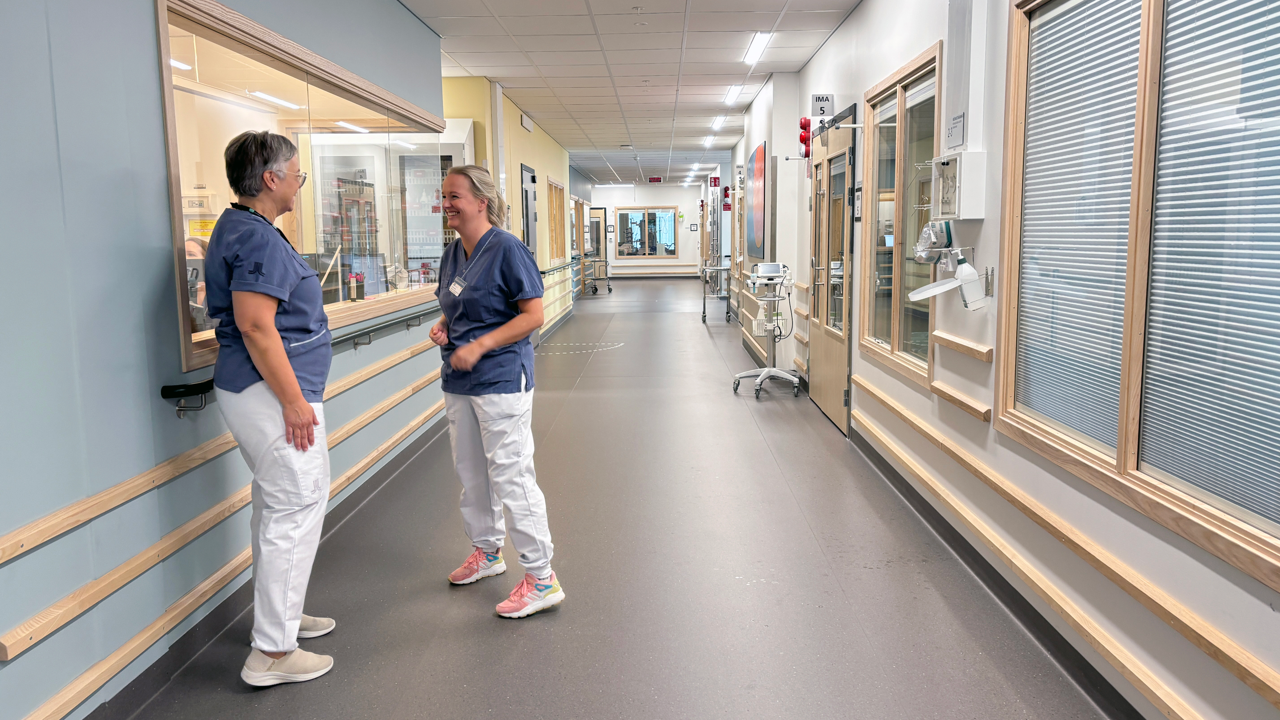 Tv&aring; kvinnor i v&aring;rdkl&auml;der st&aring;r och pratar i en korridor i v&aring;rdmilj&ouml;.