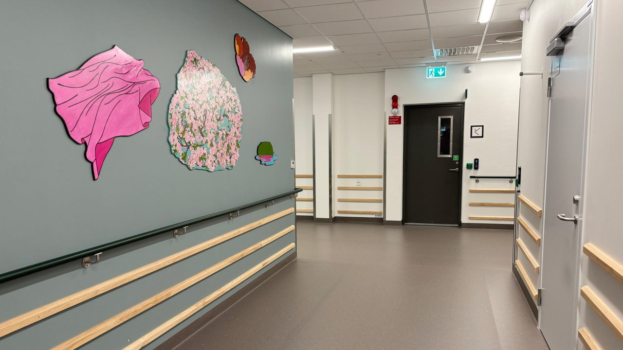 Korridor i v&aring;rdmilj&ouml; med gr&aring; matta, gr&ouml;nbl&aring; v&auml;ggar och f&auml;rgglad konst p&aring; v&auml;gg.