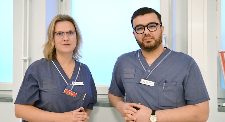 En kvinna och man i vårdkläder står vid ett fönster.