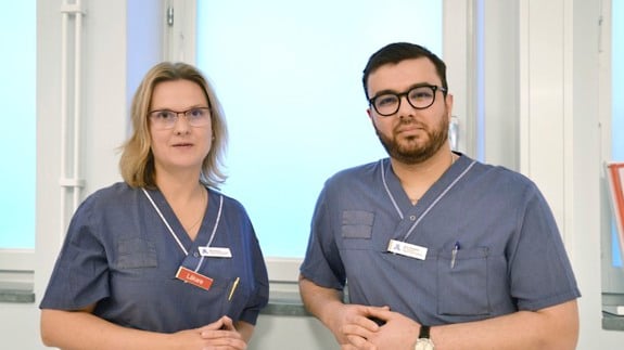 En kvinna och man i vårdkläder står vid ett fönster.