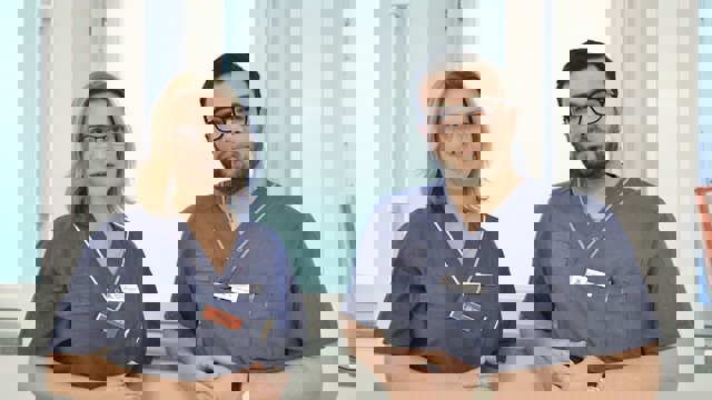 En kvinna och man i vårdkläder står vid ett fönster.