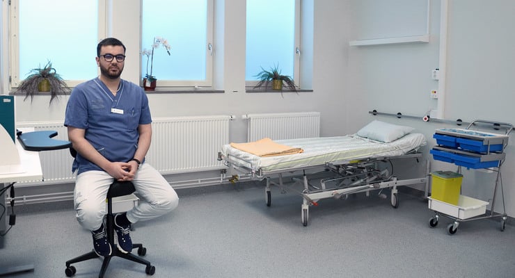 En manlig vårdpersonal sitter på en stol i ett mottagningsrum med sjukhussäng.