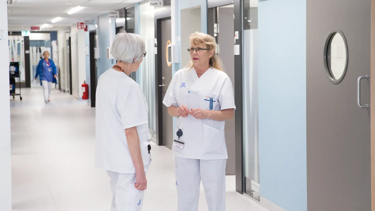Tv&aring; kvinnor i v&aring;rdkl&auml;der st&aring;r i en v&aring;rkorridor och pratar.