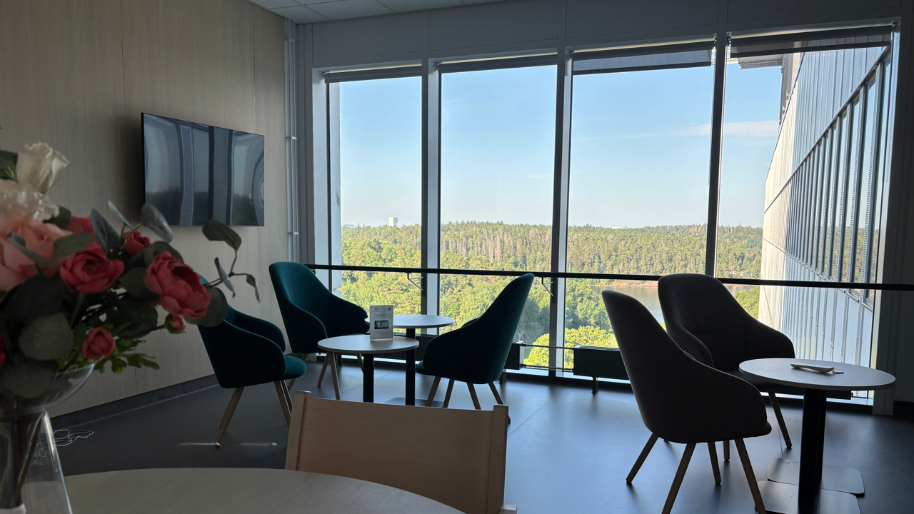 Fot&ouml;ljer och bord framf&ouml;r ett stort f&ouml;nster med utsikt mot gr&ouml;nska, vit fasad och Edsviken. 