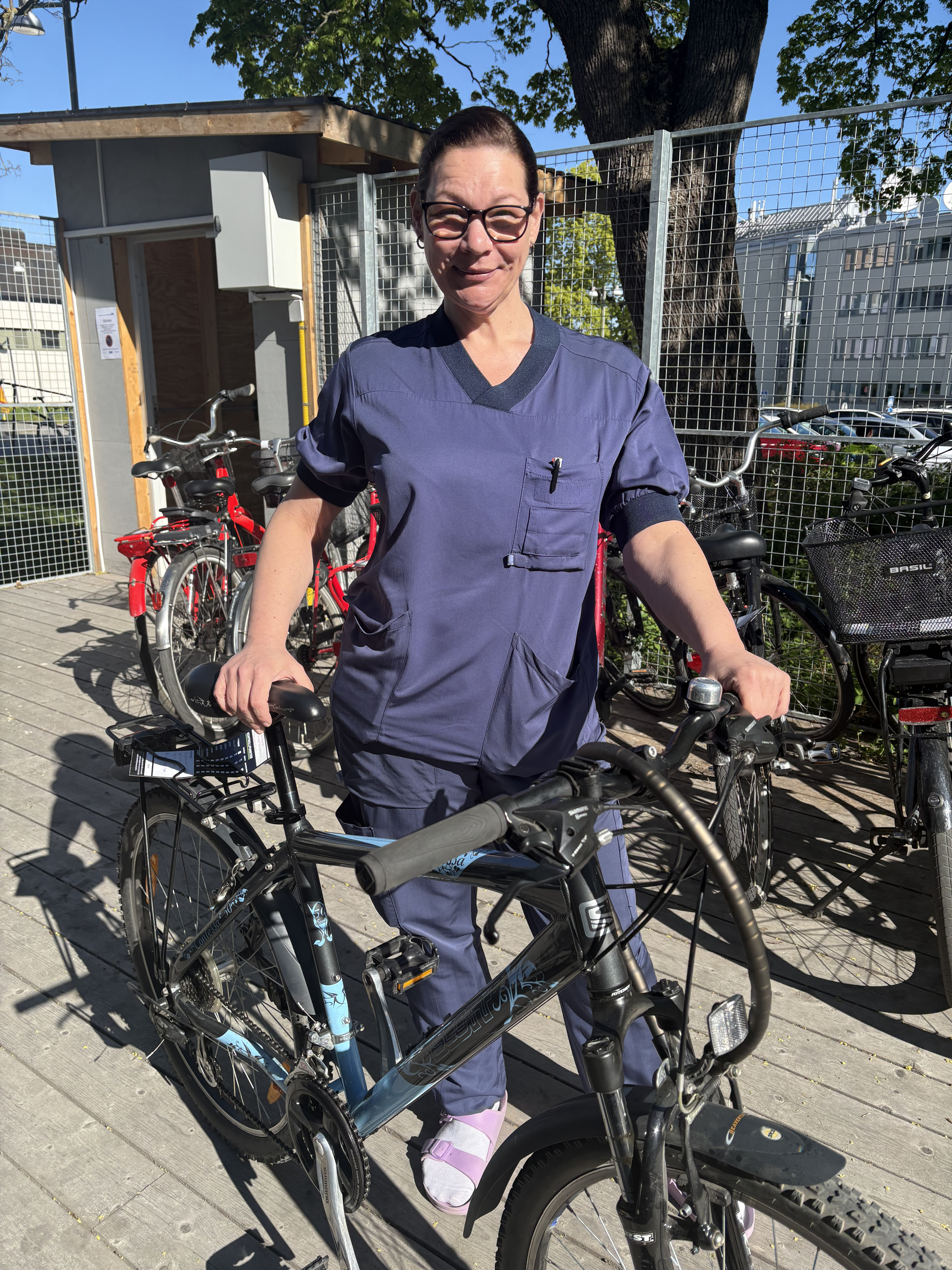 En kvinna i blå sjukhuskläder står med sin cykel och tittar in i kameran.