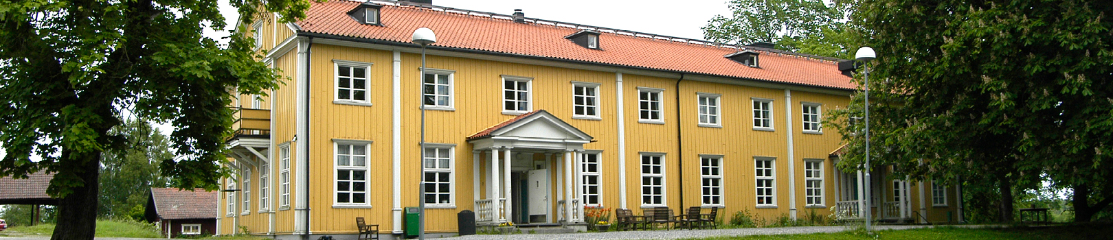 Gult trähus med rött tegeltak och vita fönsterkarmar omringad av gräsmatta och gröna stora träd.
