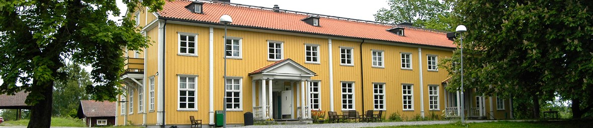 Gult trähus med rött tegeltak och vita fönsterkarmar omringad av gräsmatta och gröna stora träd.