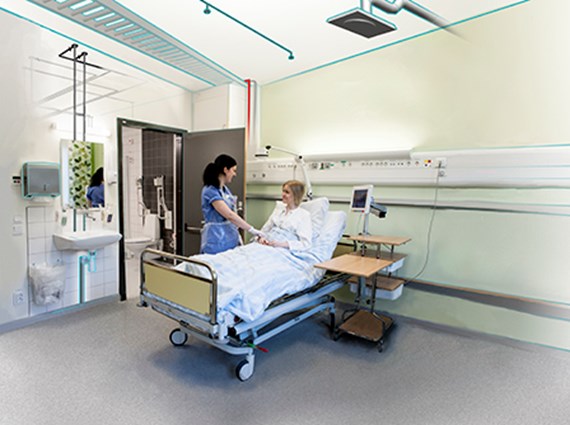 Kvinnlig vårdpersonal står vid patient som ligger i en sjukhussäng inne på ett vårdrum.