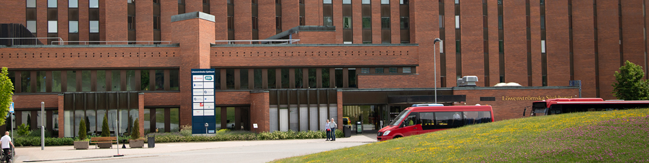 Entré till Löwenströmska sjukhusets röda tegelbyggnad i en miljö med gröna växter och träd. Röd SL-buss till höger. 