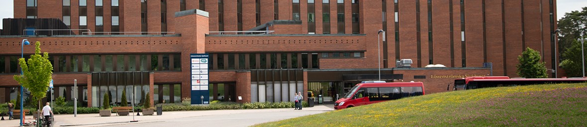 Entré till Löwenströmska sjukhusets röda tegelbyggnad i en miljö med gröna växter och träd. Röd SL-buss till höger. 
