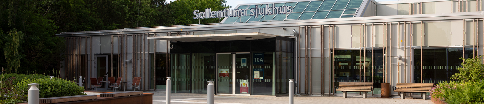 Entré till Sollentuna sjukhus gråa byggnad. Träd i bakgrunden och sittbänkar till höger om entrén.