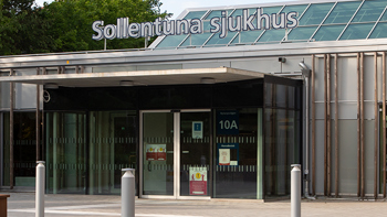 Entré till Sollentuna sjukhus gråa byggnad. Träd i bakgrunden och sittbänkar till höger om entrén.