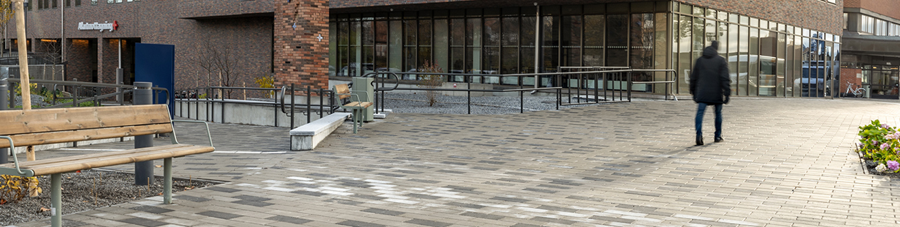 En gående suddig person går mot huvudentrén på en stenlagd gång. En parkbänk och blomplanteringar syns. Sjukhuset syns tydligt.