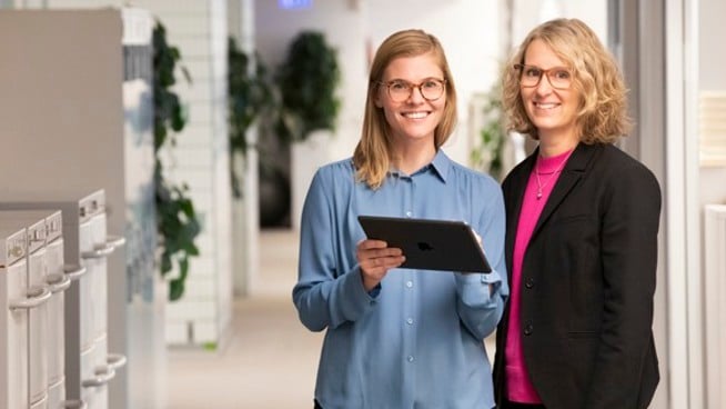 Två medarbetare på Locum står bredvid varandra i en korridor och håller i en Ipad och tittar mot oss. 