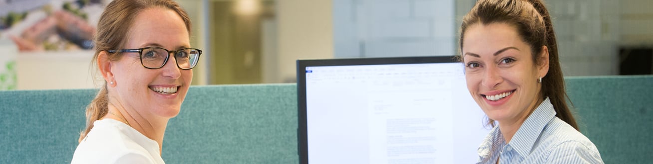 Bild på två kvinnor som står vända mot kameran framför en dataskärm