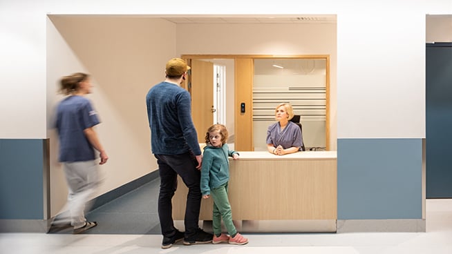 Far och dotter står vid receptionen på ett sjukhus och pratar med vårdpersonal.