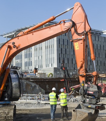 Två män i varseljackor på en byggarbetsplats, där en grävmaskin har en framträdande roll.