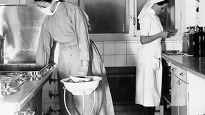 Historiskt svartvitt foto av två vårdklädda kvinnor i sjukhusmiljö.