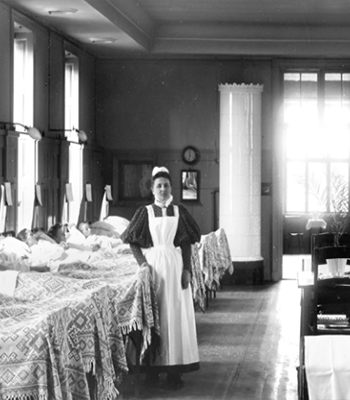 Historiskt svartvitt foto av en vårdklädd kvinna i en stor patientsal med många sängar.